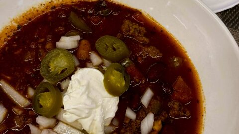 Bowl of chili topped with sour cream, jalapeños, and onions. Side plate of homemade cornbread on top of an orange towel.