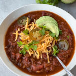 Bowl full of chili topped with shredded cheese, avocado, jalapeños, and parsley.