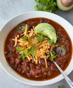 Bowl full of chili topped with shredded cheese, avocado, jalapeños, and parsley.