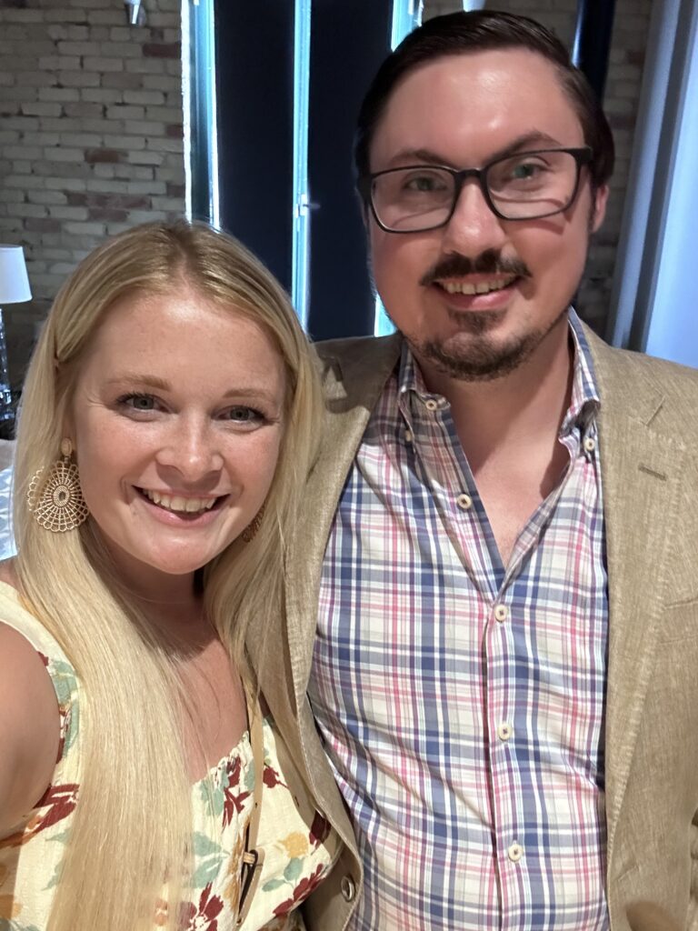 Girl wearing yellow dress and gold earrings with blonde hair standing next to a guy with glasses wearing a pink and blue plaid shirt.