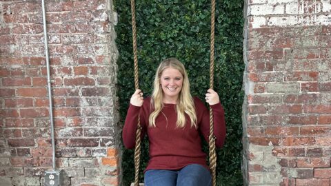 girl sitting on swing in jeans and a red sweater in front of a green ivy brick wall