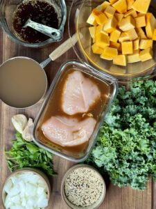 A bowl of a brown sauce, a bowl with cubed butternut squash, a bowl of marinating chicken breasts, pile of kale, bowl of dry quinoa, bowl on chopped onions, two garlic cloves, fresh parsley, and a bowl of chicken broth all on top of a wooden board.