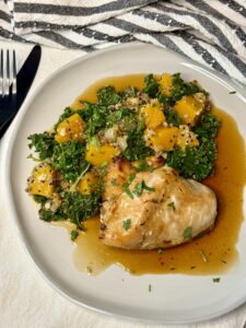 White plate with a baked chicken breast next to a pilaf of butternut squash, kale, and quinoa and a brown sauce underneath.