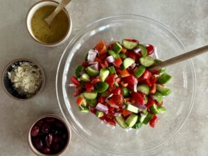 bowl full of cucumbers, tomatoes, red onion, bell pepper, olives, and feta crumbles in front of a larger bowl full of the same ingredients.