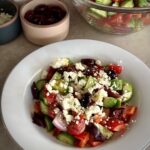 Greek salad: bowl full of cucumbers, tomatoes, red onion, bell pepper, olives, and feta crumbles in front of a larger bowl full of the same ingredients.