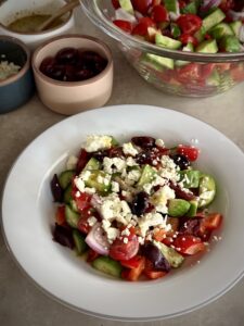 Greek salad: bowl full of cucumbers, tomatoes, red onion, bell pepper, olives, and feta crumbles in front of a larger bowl full of the same ingredients.