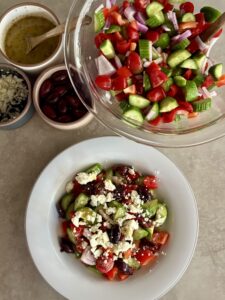 bowl full of cucumbers, tomatoes, red onion, bell pepper, olives, and feta crumbles in front of a larger bowl full of the same ingredients.