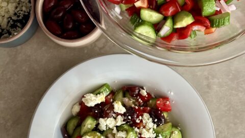 greek salad: bowl full of cucumbers, tomatoes, red onion, bell pepper, olives, and feta crumbles in front of a larger bowl full of the same ingredients.