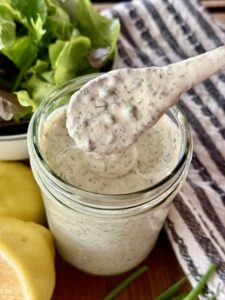 mason jar full of homemade ranch sitting on a wooden board next to fish lemon, chives, and a bowl of lettuce.