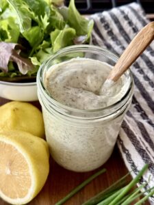 mason jar full of homemade ranch sitting on a wooden board next to fish lemon, chives, and a bowl of lettuce.