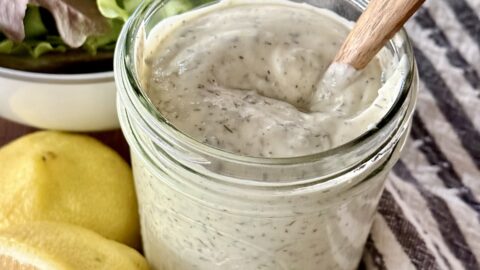 mason jar full of homemade ranch sitting on a wooden board next to fish lemon, chives, and a bowl of lettuce.