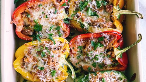 colorful bell peppers cut in half laying in a baking pan. stuffed with sausage mixture and topped with cheese.