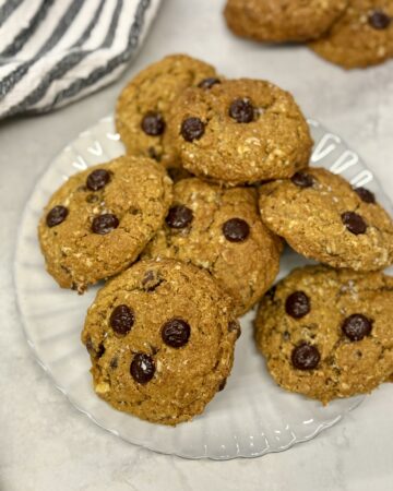 baked pumpkin oatmeal chocolate chip cookies piled on a grey scalloped plate with two cookies in the background