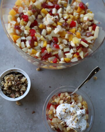 giant glass bowl full of small diced fruit, side bowl of chopped walnuts, and a bowl of the fruit salad topped with fresh whipped cream.