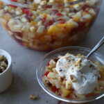 close up of a giant glass bowl full of small diced fruit, side bowl of chopped walnuts, and a bowl of the fruit salad topped with fresh whipped cream.