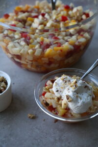 close up of a giant glass bowl full of small diced fruit, side bowl of chopped walnuts, and a bowl of the fruit salad topped with fresh whipped cream.