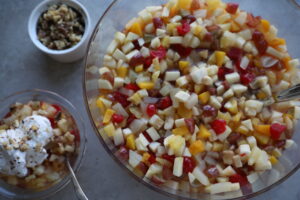 giant glass bowl full of small diced fruit, side bowl of chopped walnuts, and a bowl of the fruit salad topped with fresh whipped cream.