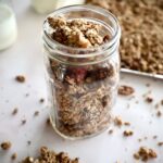 Homemade granola in a glass jar. Behind the glass jar is more granola on a baking sheet, and to the left are two glass jars with milk.