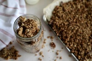 Homemade granola in a glass jar. Behind the glass jar is more granola on a baking sheet, and to the left are two glass jars with milk.