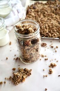 Homemade granola in a glass jar. Behind the glass jar is more granola on a baking sheet, and to the left are two glass jars with milk.