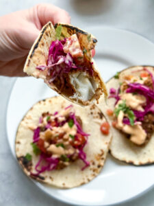 plate of three tacos shells topped with crispy baked fish tacos. next to the plate is a bowl of the creamy chipotle sauce, cabbage slaw, and cut-up limes.