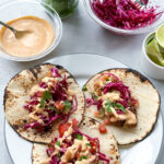 plate of three tacos shells topped with crispy baked fish tacos. next to the plate is a bowl of the creamy chipotle sauce, cabbage slaw, and cut-up limes.