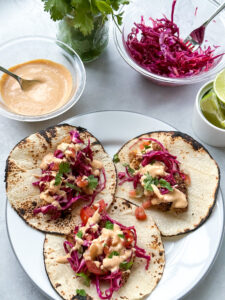 plate of three tacos shells topped with crispy baked fish tacos. next to the plate is a bowl of the creamy chipotle sauce, cabbage slaw, and cut-up limes.