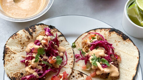 plate of three tacos shells topped with crispy baked fish tacos. next to the plate is a bowl of the creamy chipotle sauce, cabbage slaw, and cut-up limes.