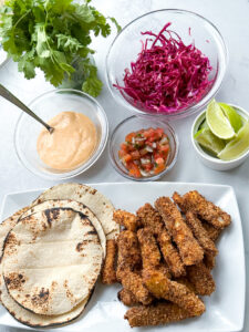 plate of three tacos shells topped with crispy baked fish tacos. next to the plate is a bowl of the creamy chipotle sauce, cabbage slaw, and cut-up limes.